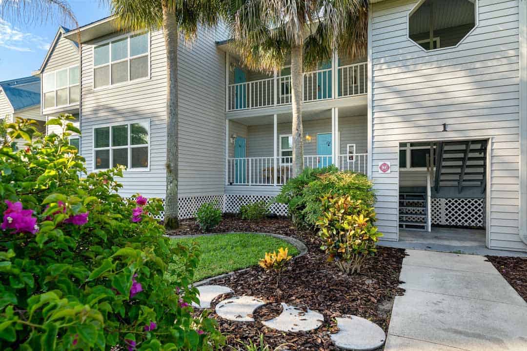 A residential building with three stories and balconies, surrounded by lush landscaping and palm trees, under a clear blue sky.
