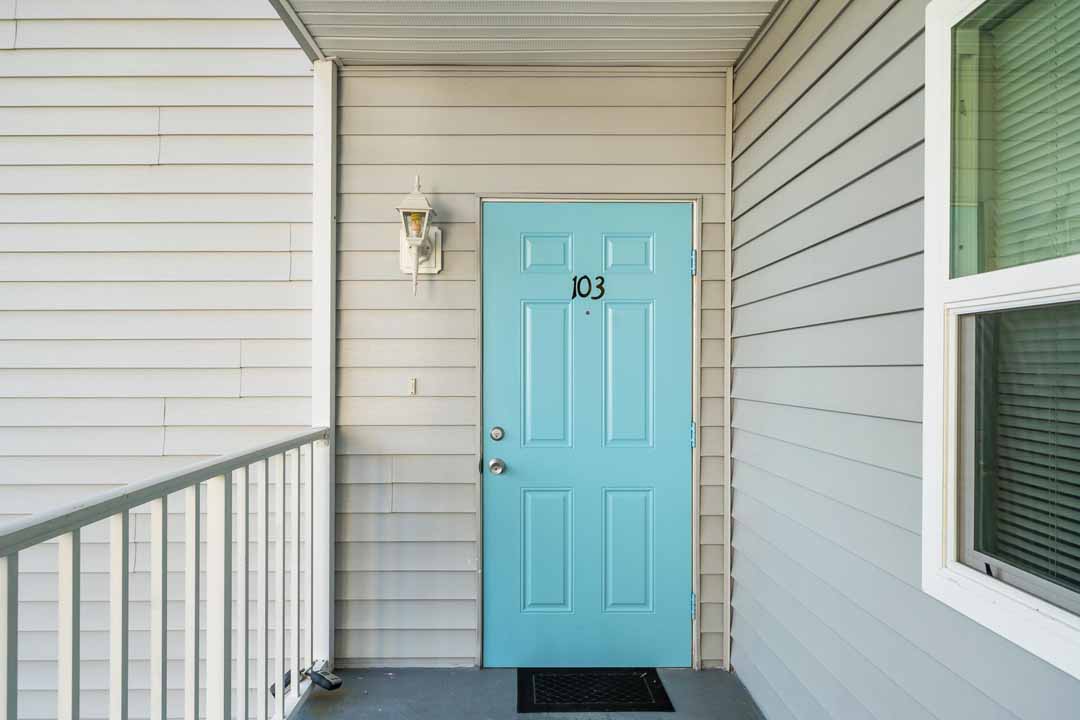 Light blue door numbered 103 on a residential building with white siding, featuring a porch, railing, and a window to the right.