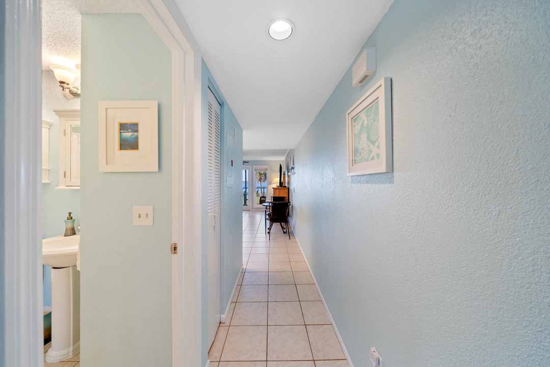 Narrow hallway in a home with light blue walls, tiled floor, framed pictures, and a view into a room with a dining table at the end.