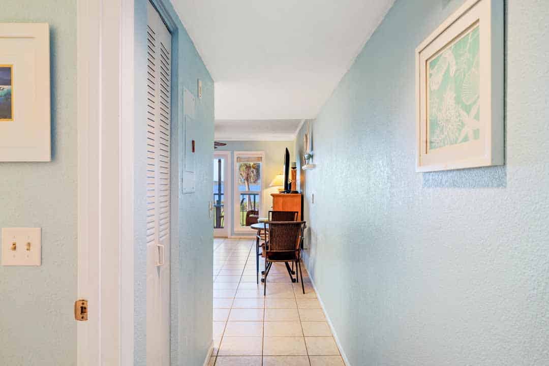 Brightly lit hallway in a house with teal walls, framed artwork, and a wooden chair at the end.