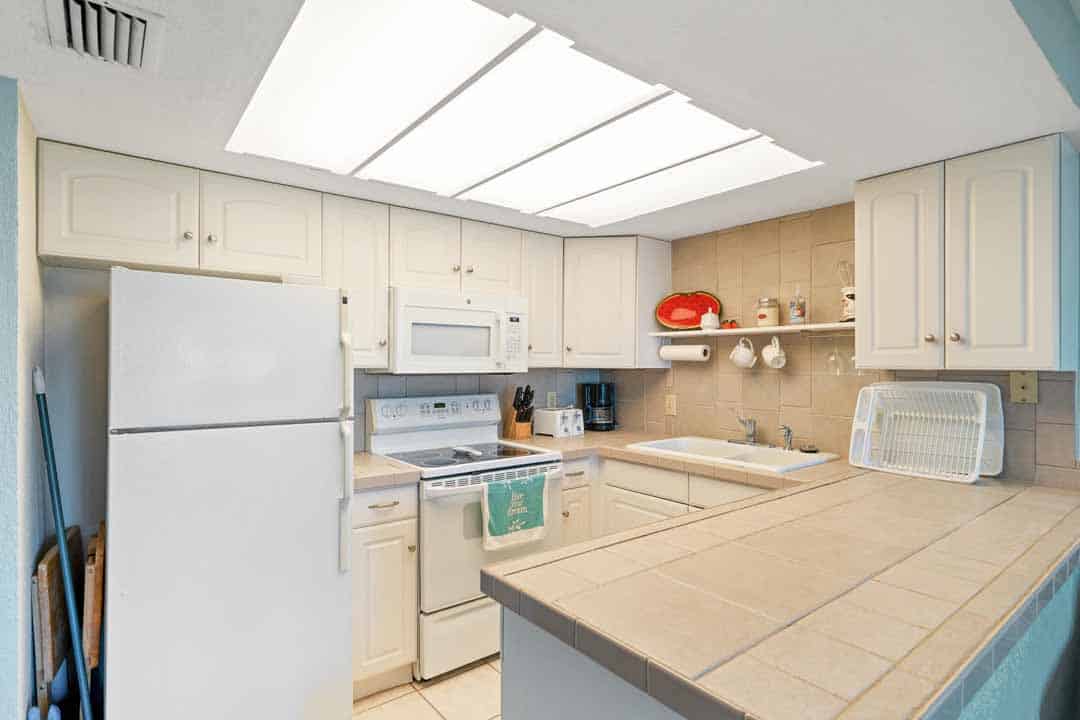 A bright, compact kitchen with white cabinets, appliances, and a skylight, showing a microwave, stove, fridge, and a sink with a drying rack.