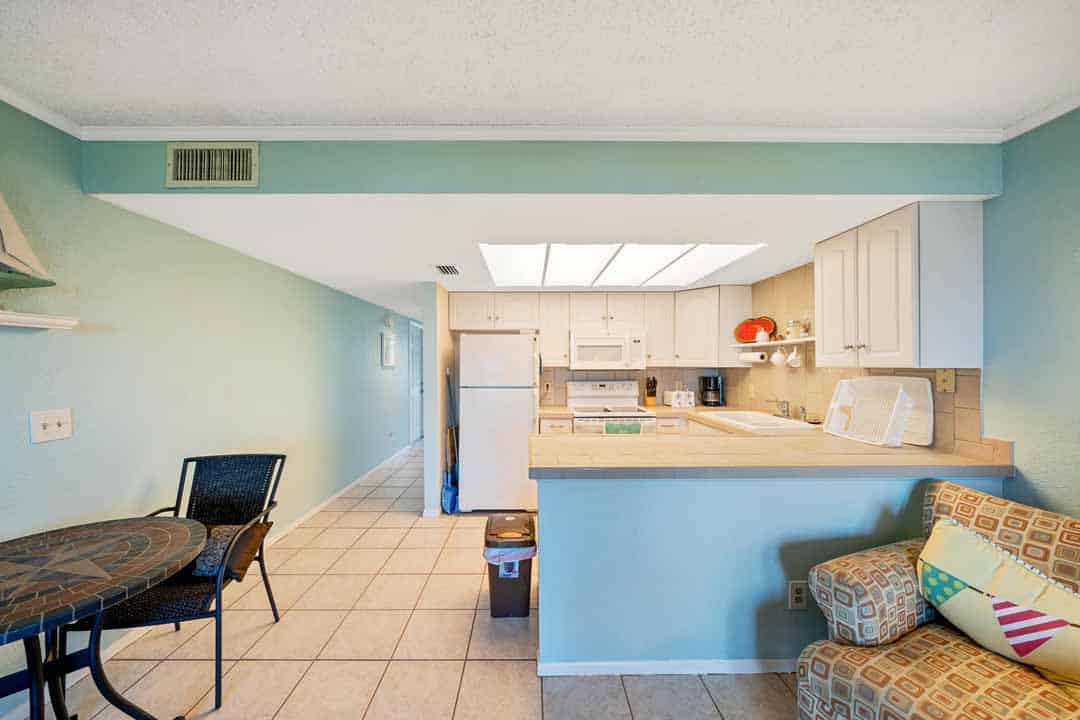 Bright, compact kitchen with white cabinetry and appliances, viewed from an adjoining dining area with a table and cushioned chairs.