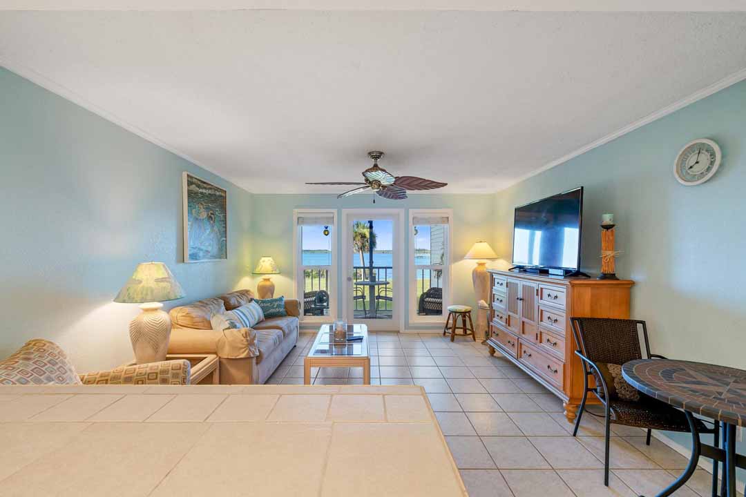Brightly lit living room with a beige sofa, armchairs, tv, and a dining area leading to a balcony with an ocean view.