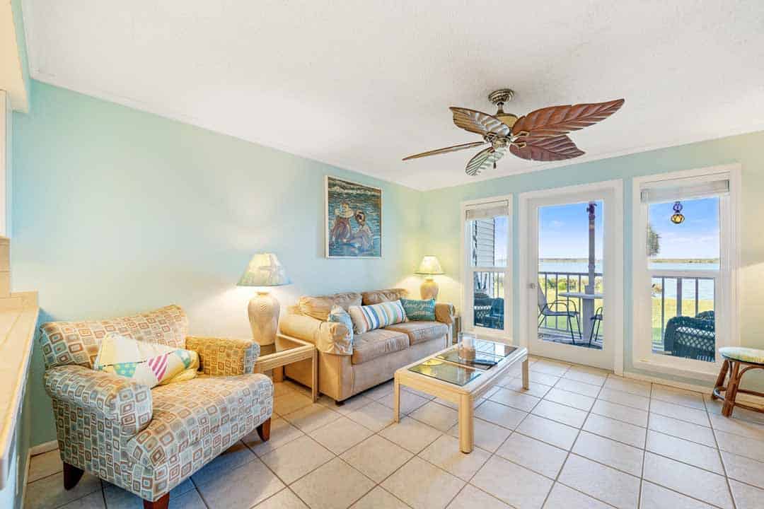 Brightly lit beachfront living room with couch, armchair, ceiling fan, and sliding doors opening to a balcony overlooking the sea.