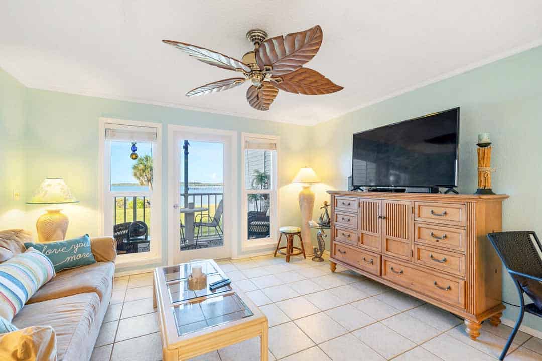 Bright, coastal-style living room with a sofa, tv, ceiling fan, and large windows opening to a beachfront balcony.