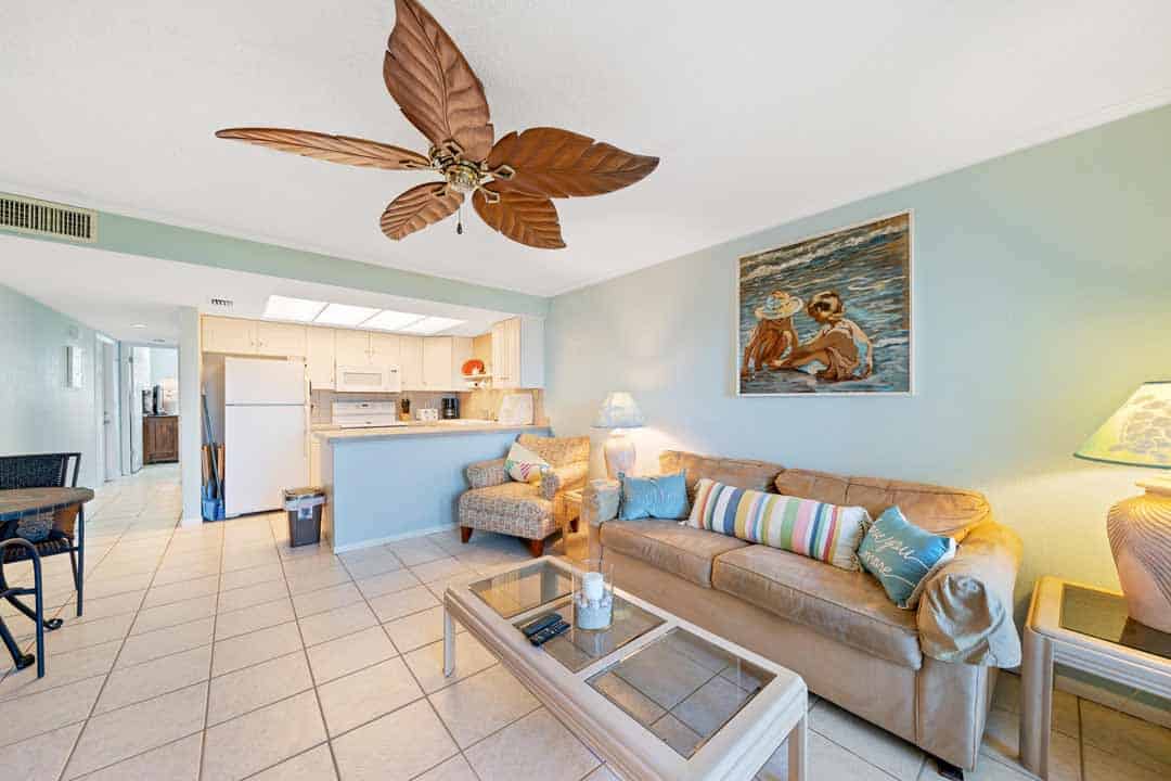 Bright, airy living room with a couch, chairs, ceiling fan, and open kitchen, featuring coastal decor and a painting of swimmers on the wall.