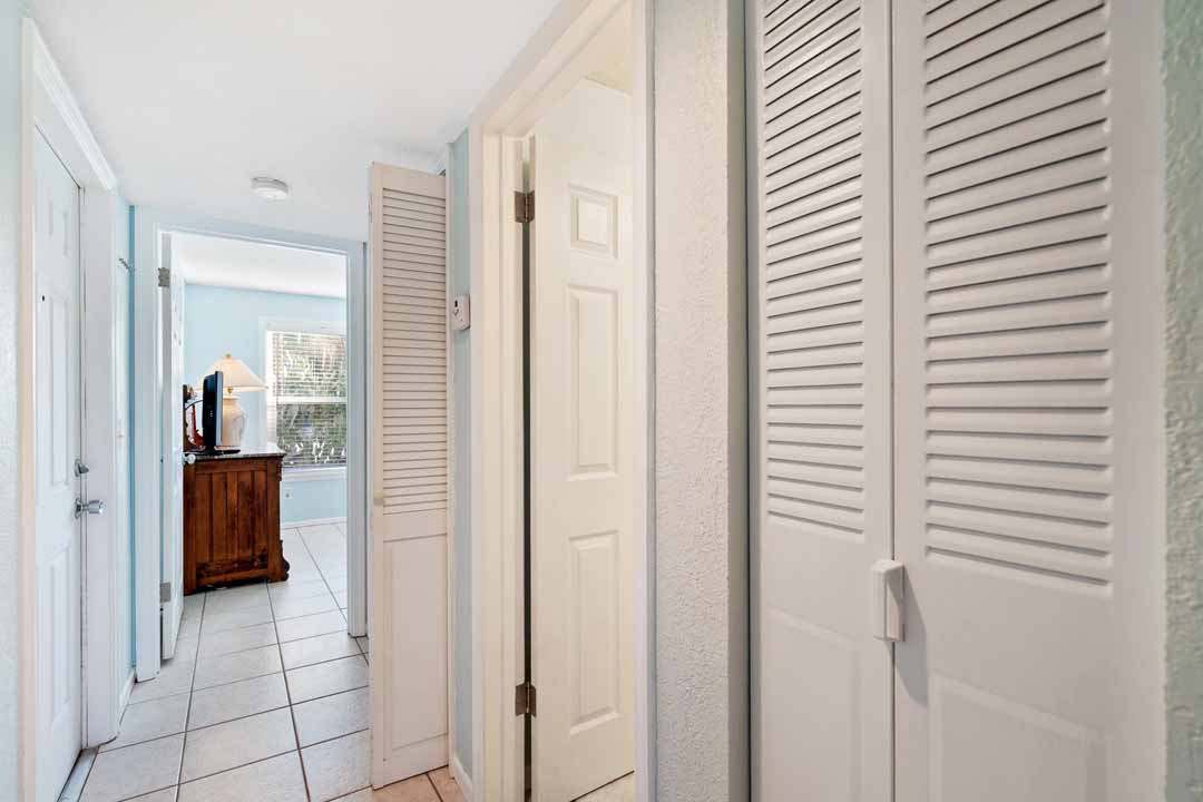 Brightly lit hallway in a modern home with white walls, louvered doors on the left, and a view into a sunlit room at the end.