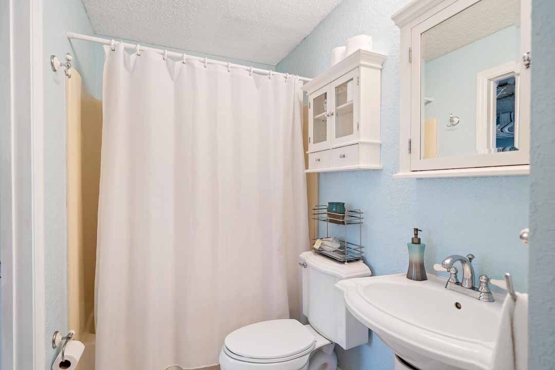 A small bathroom with pale blue walls, featuring a white shower curtain, sink, and toilet, with a white cabinet and mirror above the sink.