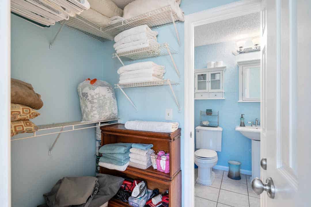 A well-organized bathroom with open shelves stocked with towels, toiletries, and a wooden cabinet containing linens, viewed from an adjacent closet.