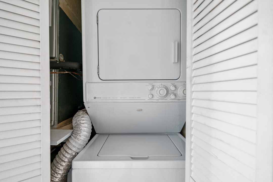 A white front-loading washing machine and dryer stacked inside a closet with louvered doors, visible piping and ductwork.