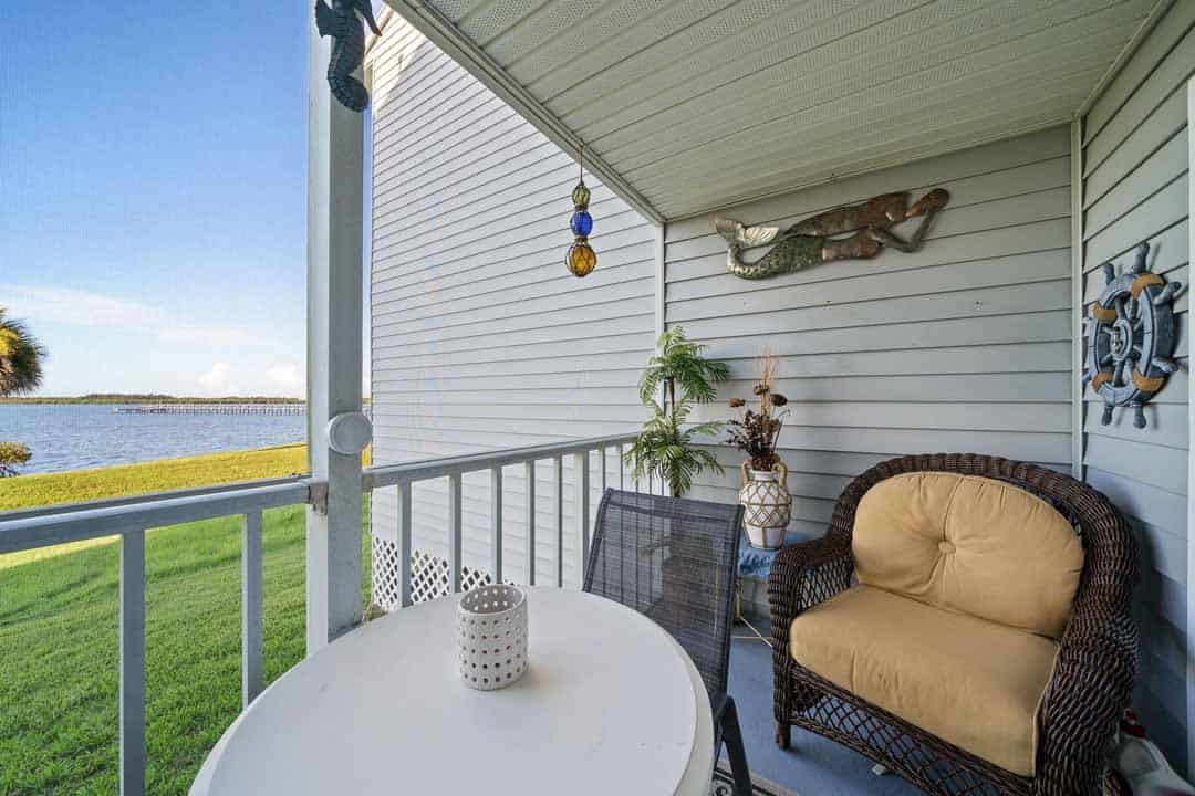 A cozy balcony with a wicker armchair and decorative items, overlooking a calm lake under a clear blue sky.