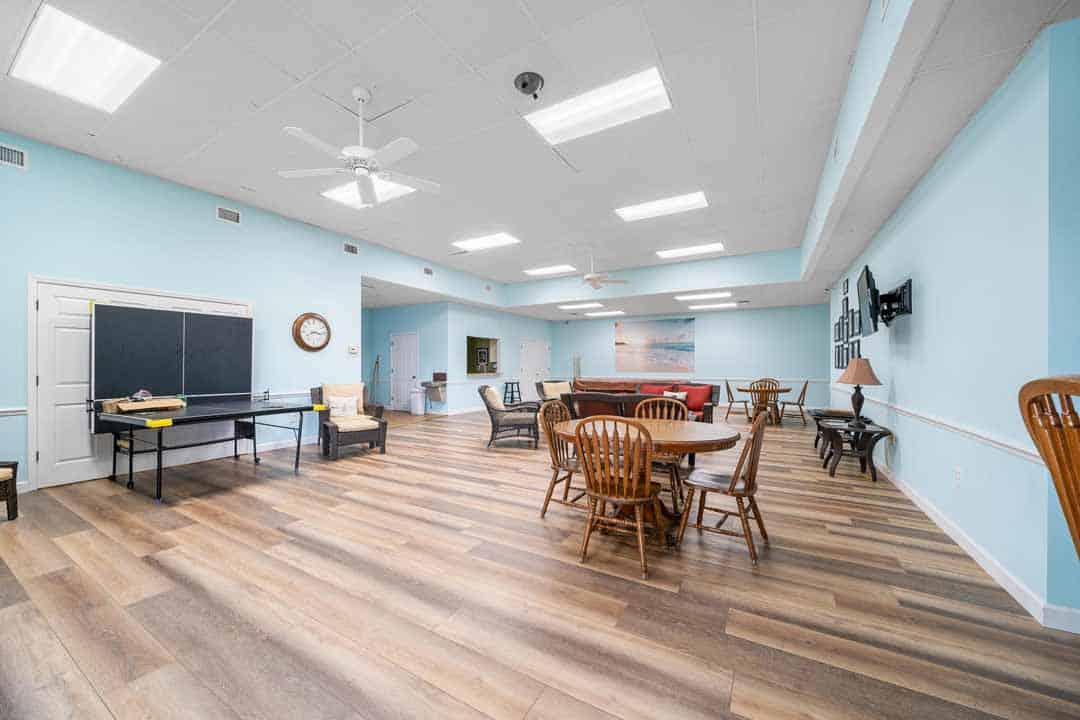 Spacious community room with wooden floors, a black chalkboard, dining tables, chairs, and a living area with sofas. decor includes wall clocks and art.