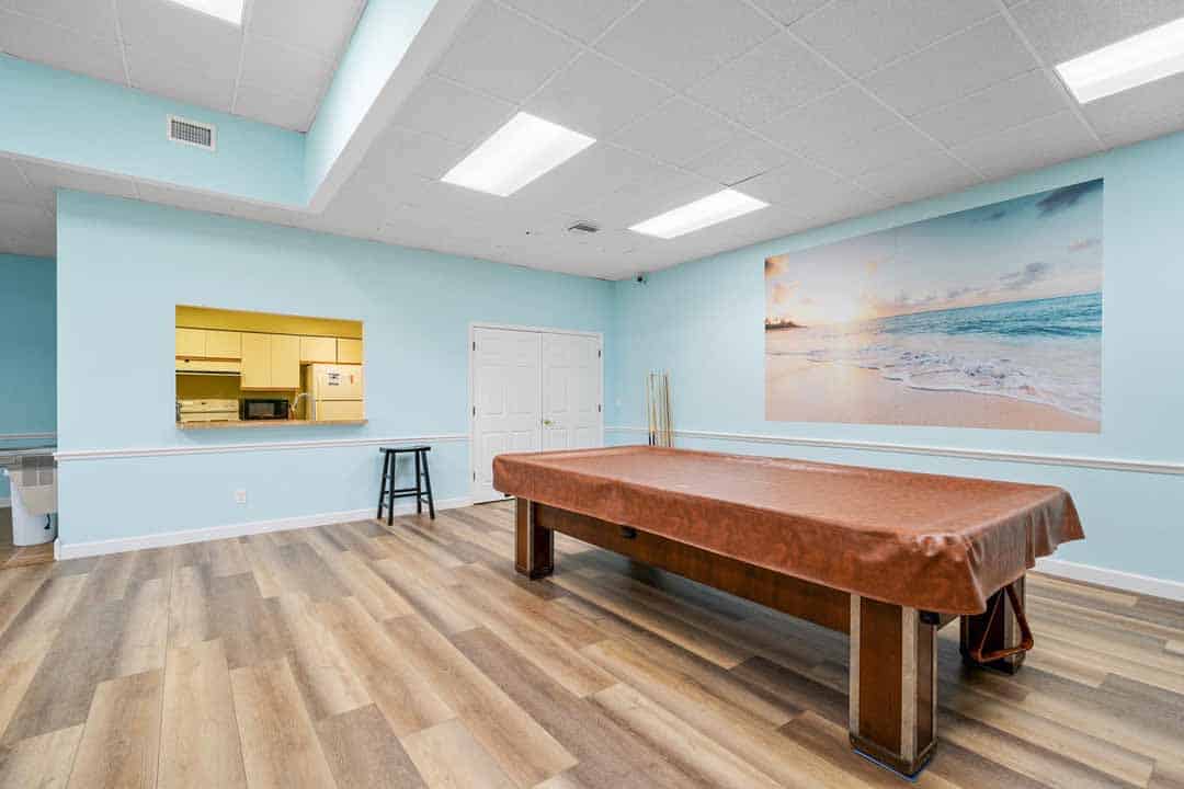 Bright room with a large table, beach mural, wood floors, and kitchenette visible through an open passage.