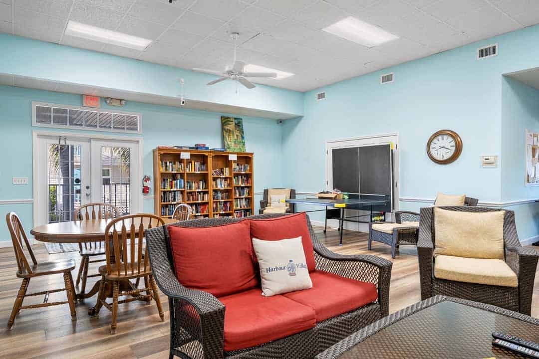 Bright community room with red sofa, chairs, bookshelves, a blackboard, and a ceiling fan, featuring light blue walls and wood flooring.
