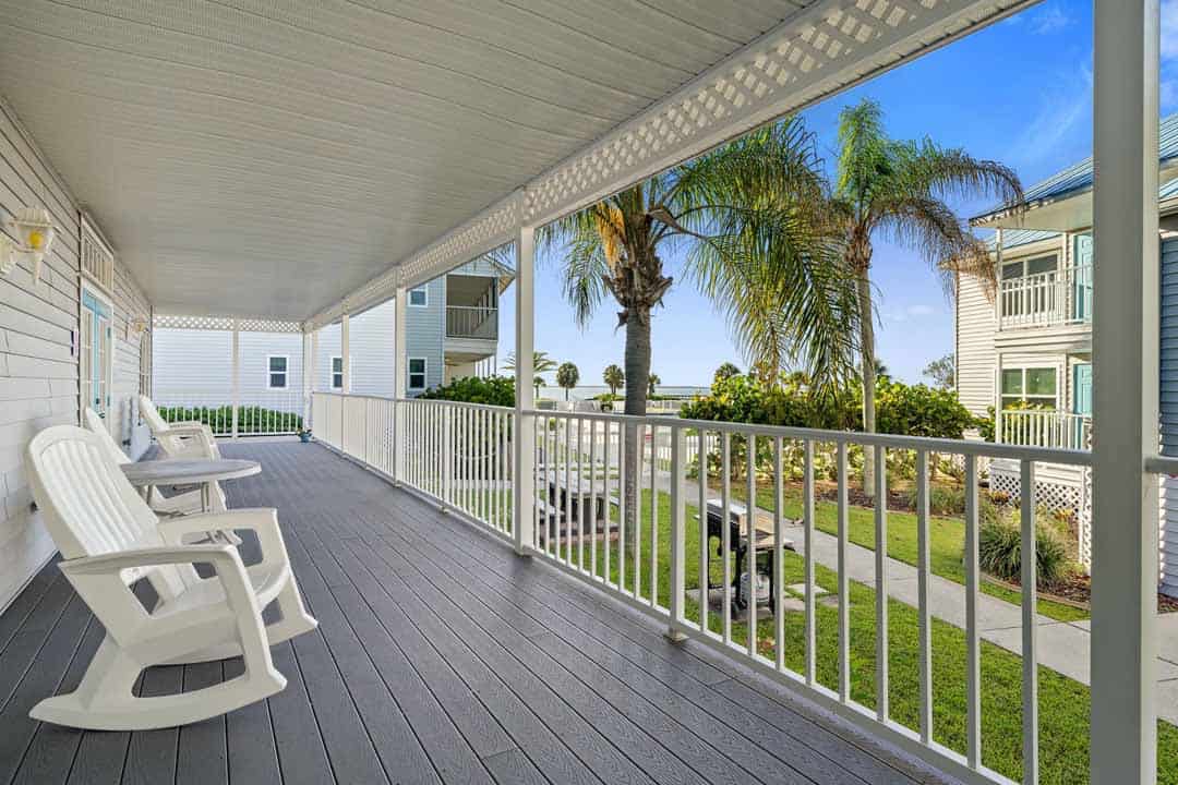 A spacious porch with white railings and flooring, featuring several chairs and overlooking surrounding tropical landscapes with palm trees.