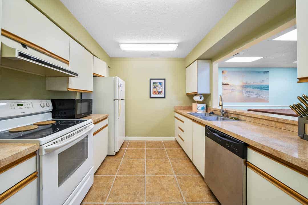 A bright, compact kitchen with beige cabinets, granite countertops, and modern appliances, featuring a microwave, stove, and dishwasher, with a beach-themed picture on the wall.