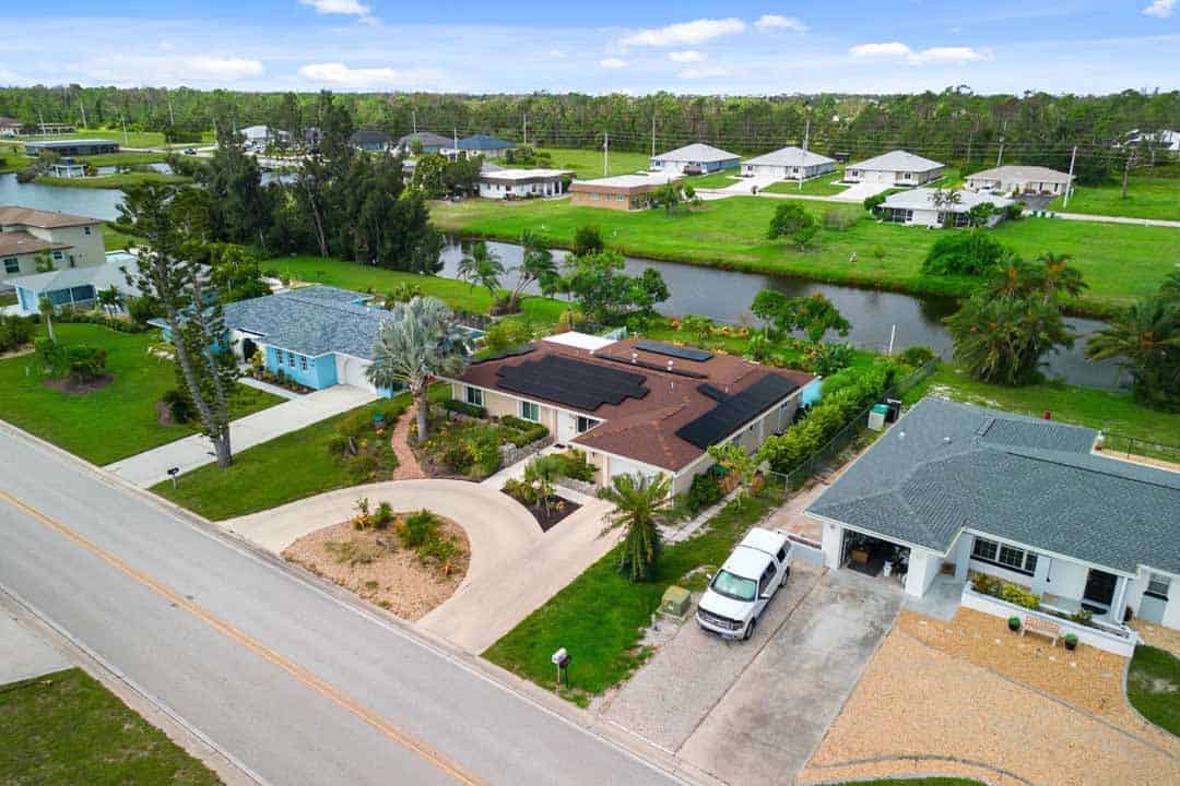 Aerial view of a residential Rotonda neighborhood with modern homes alongside a road and a canal, featuring lush greenery and parked cars.