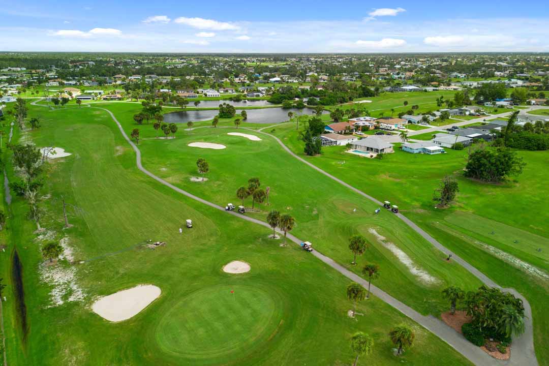 Aerial view of a golf course in the Rotonda community, with green fairways, sand traps, and golf carts, surrounded by residential areas under a clear blue sky.