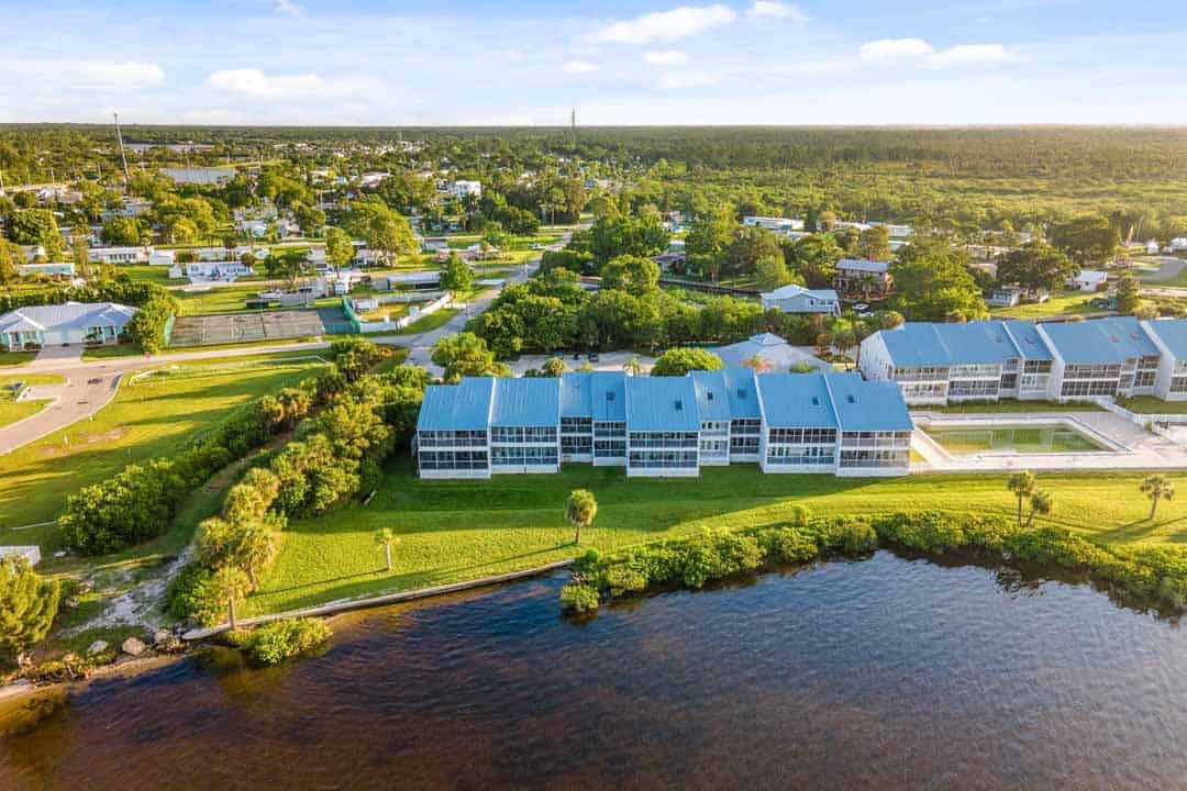 Aerial view of a riverside residential area with clustered white buildings surrounded by green spaces and trees under a clear sky.