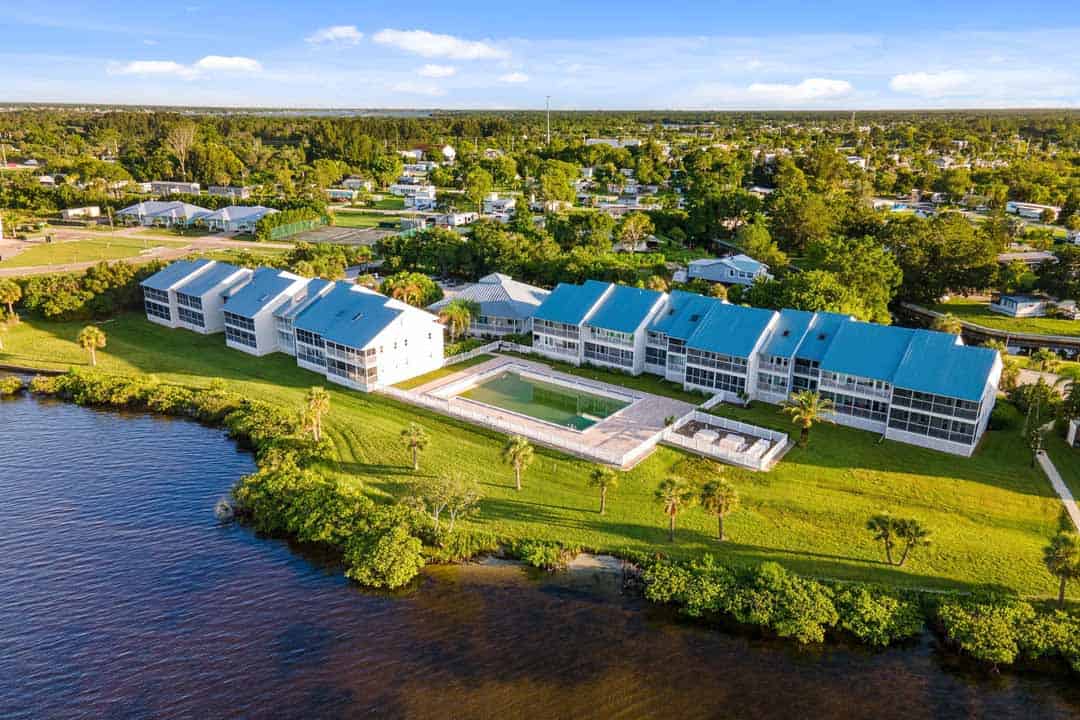 Aerial view of a modern residential complex with blue roofs beside a river, surrounded by lush greenery in a suburban setting.