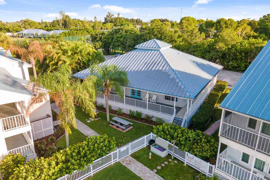 Aerial view of a suburban home with a blue metal roof surrounded by lush greenery and neighboring houses.