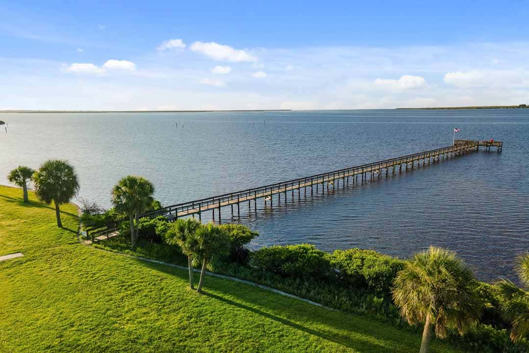 Aerial view of a long wooden pier extending into a large body of water, surrounded by green grass and palms under a clear blue sky.