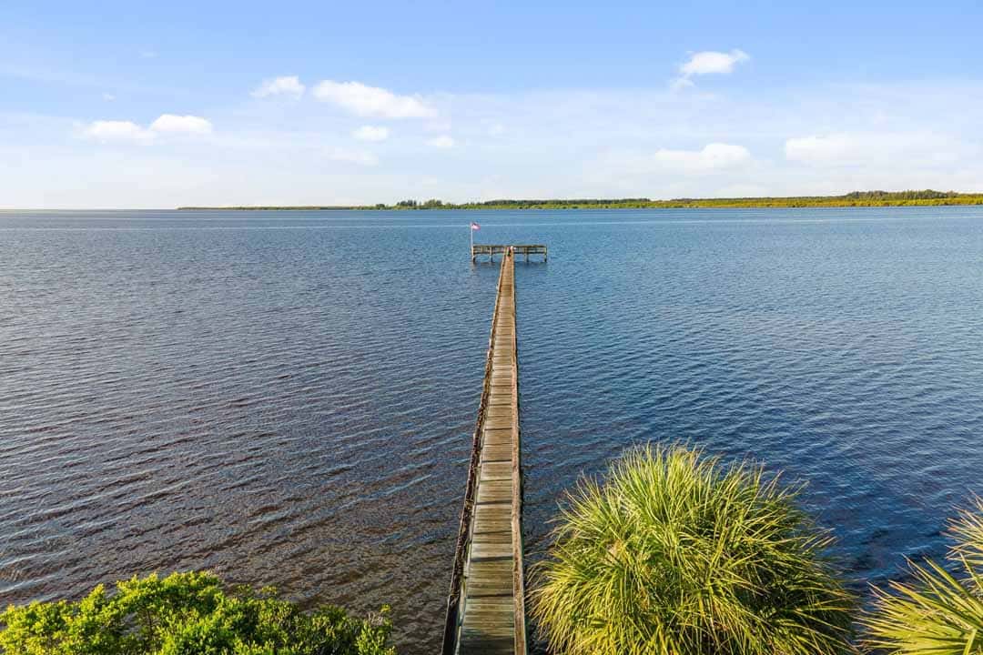 A wooden pier extends into a calm lake, surrounded by lush greenery under a clear blue sky.