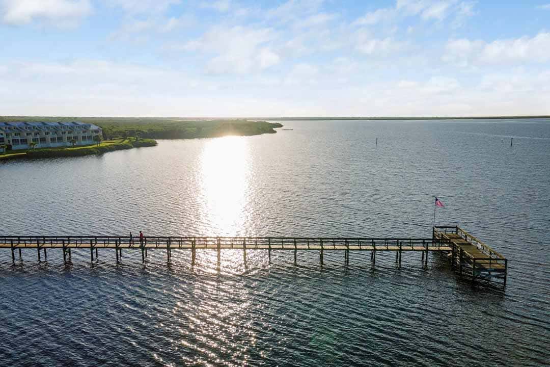Aerial view of a wooden pier extending into a calm bay at sunrise, with a person standing near the end and coastal buildings in the background.