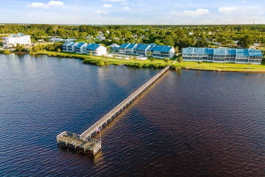 Aerial view of a long wooden pier extending into a river, with rows of blue-roofed buildings along the shore under a clear sky.
