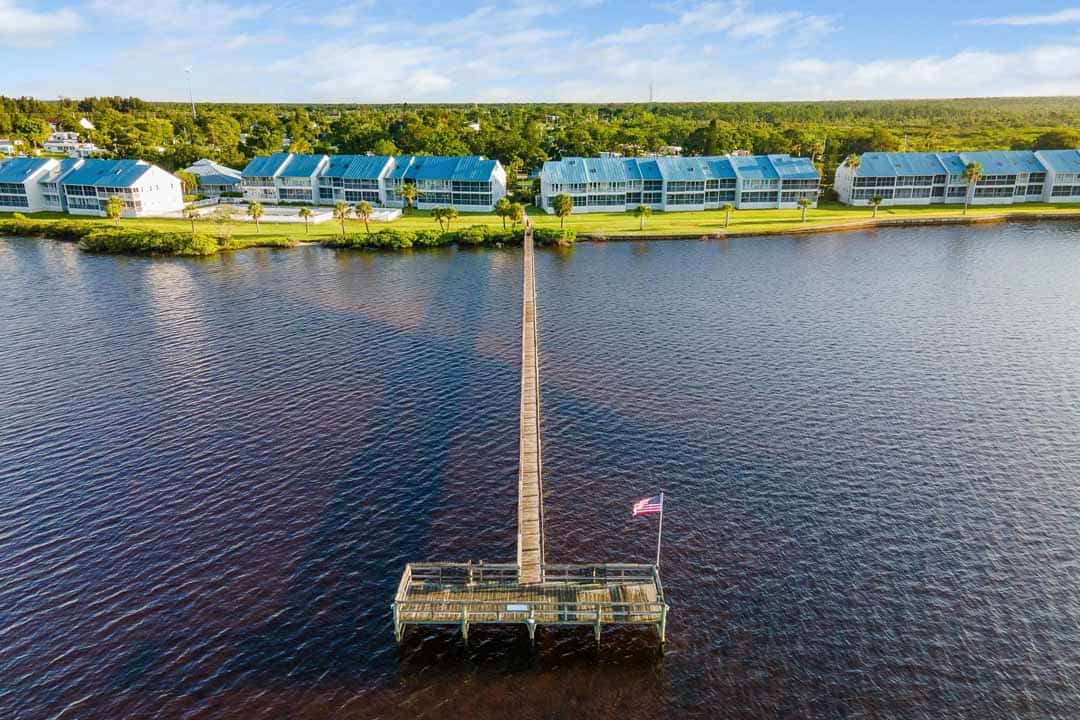 Aerial view of a row of waterfront townhouses with blue roofs, connected to a small pier flying the american flag, beside a calm river.
