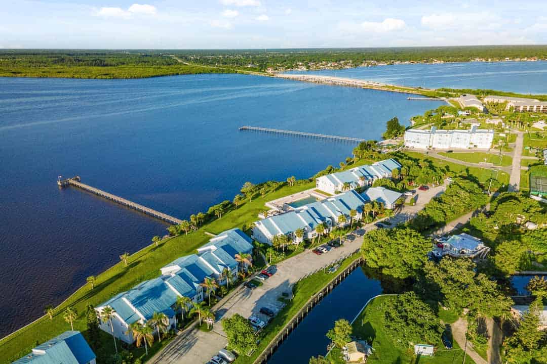 Aerial view of a coastal town with rows of light blue houses along a canal, adjacent to a broad river and greenery.