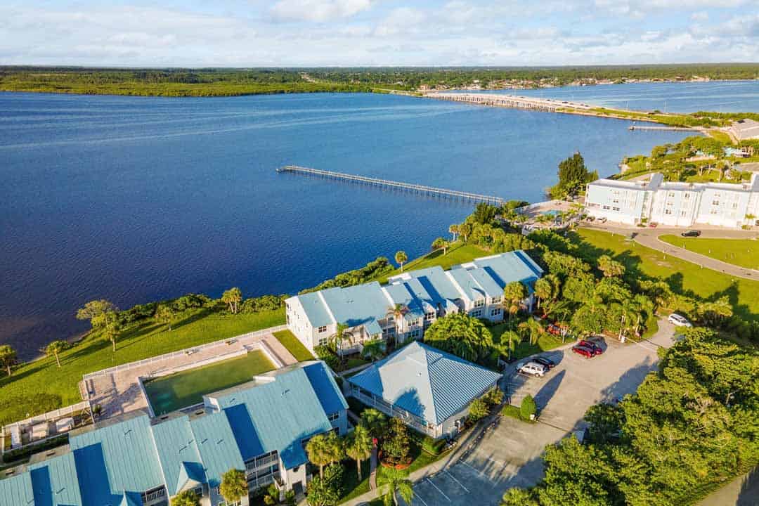 Aerial view of a coastal residential area with rows of buildings and a long pier extending into a wide river, surrounded by greenery and a clear sky.