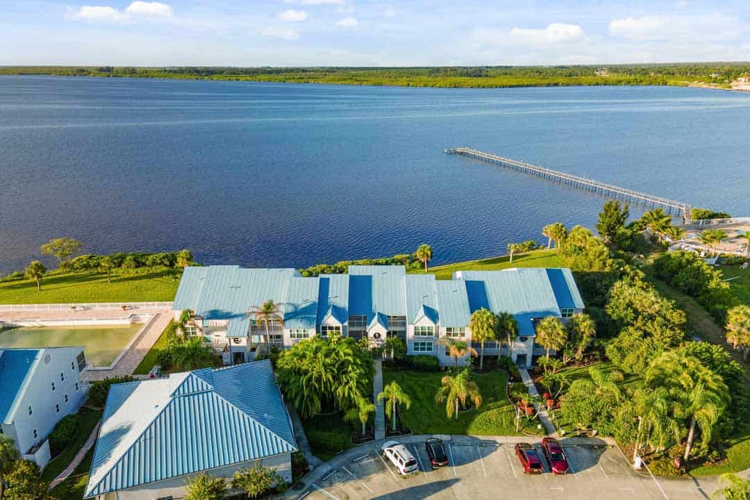 Aerial view of a waterfront property with blue-roofed buildings surrounded by lush greenery, a long pier extending into a calm lake under a clear sky.