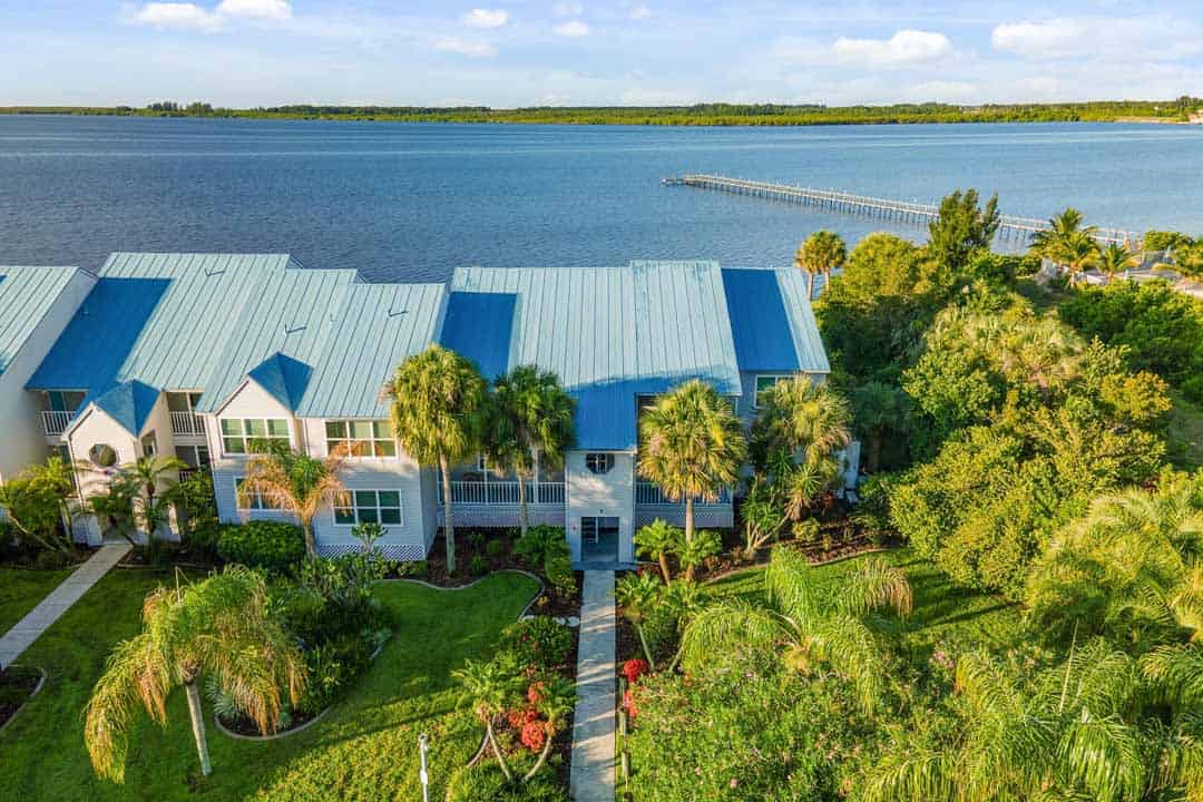 Aerial view of a coastal residential area with blue-roofed buildings, lush greenery, and a long pier extending into the calm water.