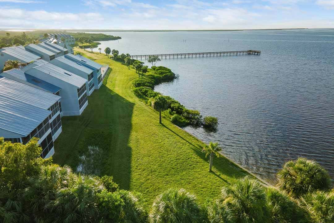 Aerial view of a coastal landscape featuring modern buildings, a lush green lawn, trees, and a long pier extending into a calm large body of water.