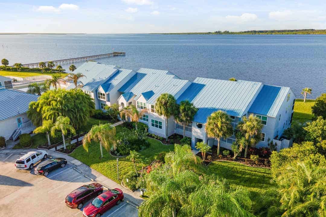 Aerial view of a coastal residential area with blue-roofed houses, parked cars, and palm trees near a large body of water with a pier extending into it.