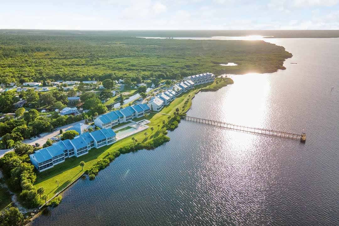 Aerial view of a coastal residential area with rows of blue-roofed buildings along a river, connected to a jetty, with lush greenery surrounding and the sun reflecting on the water.