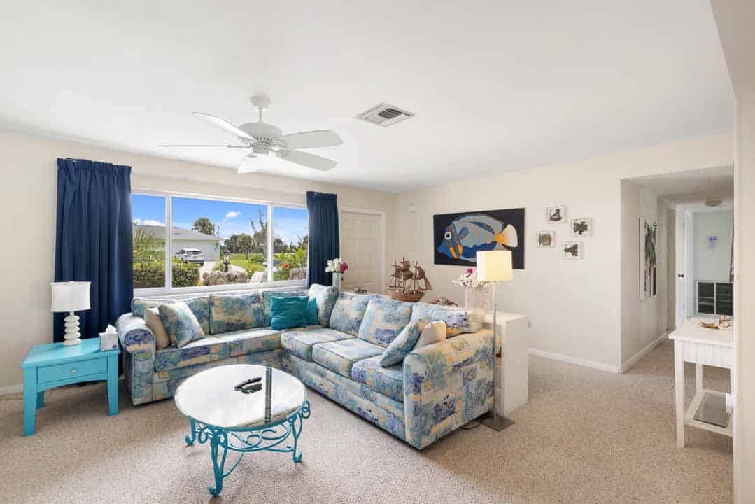 Bright, contemporary living room in a planned community, with a floral sofa, white coffee table, and blue accents, overlooking a sunny view through large windows.