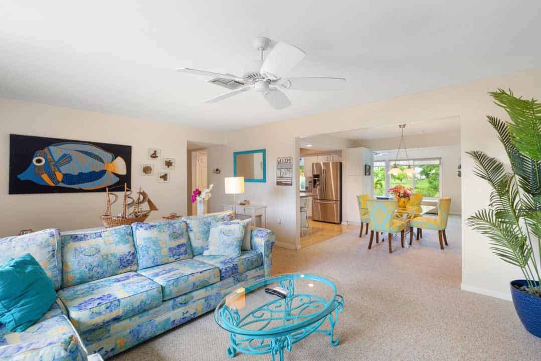 Bright, airy living room with a blue patterned sofa, circular glass table, Rotonda wall art, and ceiling fan, opening into a dining area with a yellow table.