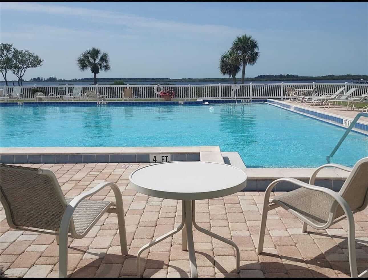 Outdoor swimming pool with a view of a lake, surrounded by a white fence. Two chairs and a table are set on a brick patio in the foreground. Palm trees and distant trees are visible.