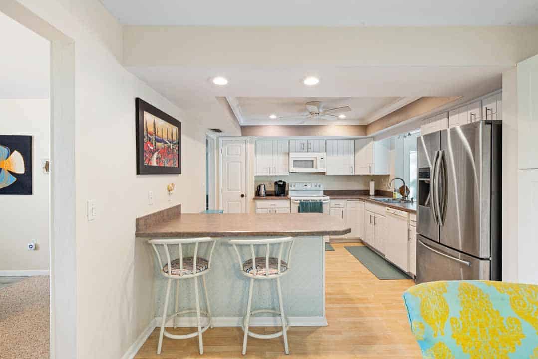 A modern kitchen with white cabinetry, stainless steel appliances, and a breakfast bar with two stools, adjoining a living area.
