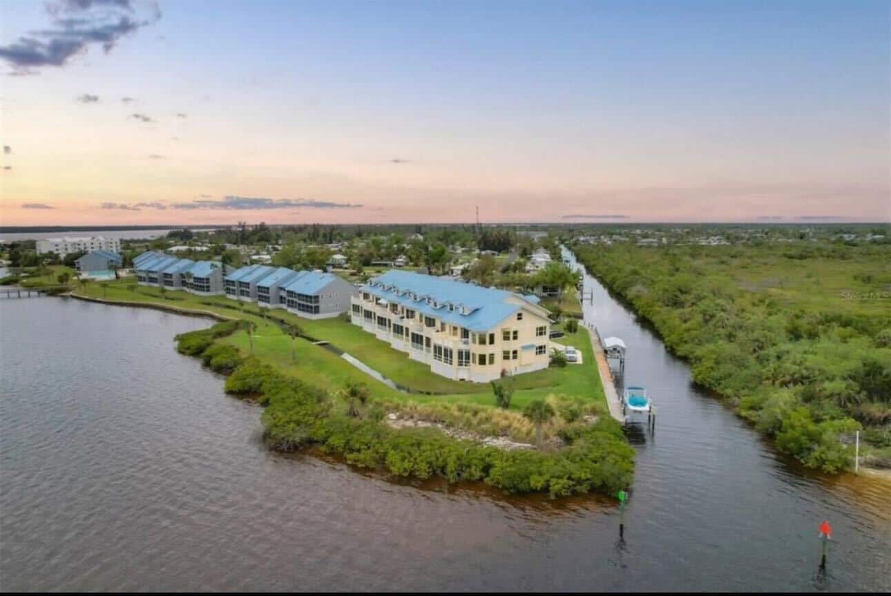 Aerial view of a waterfront complex with multiple buildings featuring blue roofs, adjacent to a canal with boats, set against a sunset sky.
