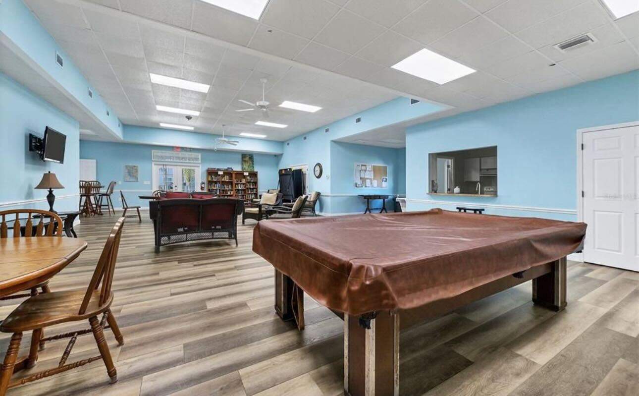 A spacious recreational room with a pool table, lounge chairs, a television, a bookshelf, tables, and chairs. The room has blue walls, wood flooring, and ceiling lights.