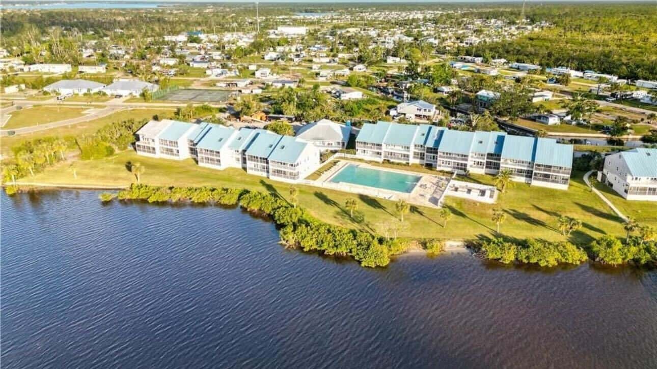 Aerial view of a residential complex with multiple buildings, a central swimming pool, and lush greenery, situated next to a body of water in a suburban area.