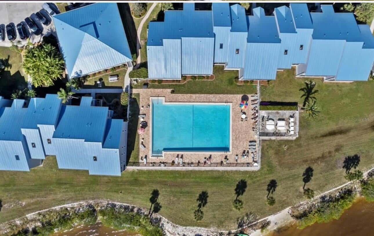 Aerial view of a rectangular swimming pool surrounded by buildings with blue roofs. Adjacent grassy area with palm trees and tables. There is a body of water at the bottom edge of the image.