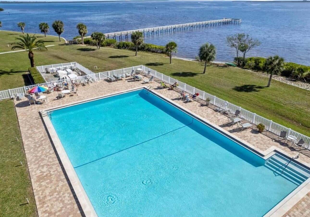 A large outdoor swimming pool with lounge chairs and umbrellas is adjacent to a grassy area and a body of water with a wooden pier extending into it. Palm trees are scattered throughout the scene.