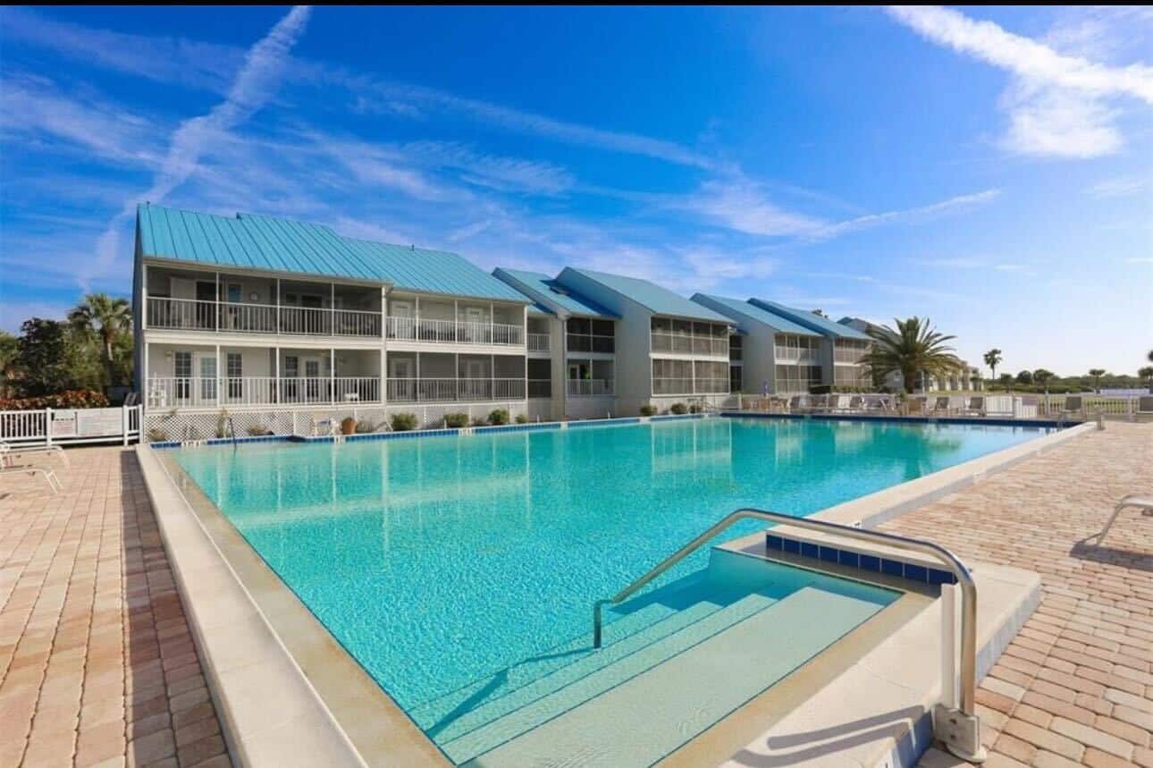 Outdoor swimming pool with clear blue water in front of a two-story modern building with blue roofs and balconies under a bright blue sky.