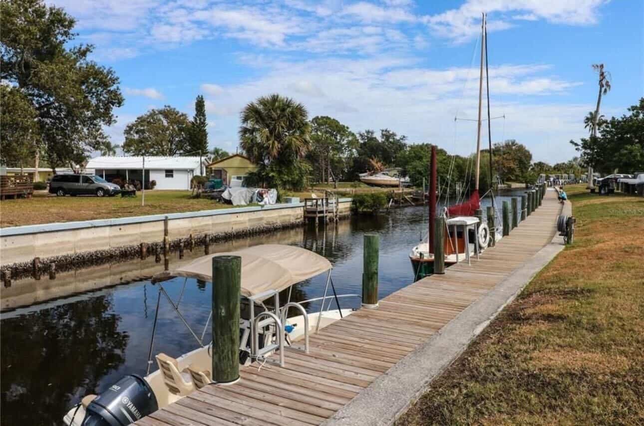 A small canal lined with boats docked at a wooden pier, with houses, trees, and grass on either side under a partly cloudy sky.