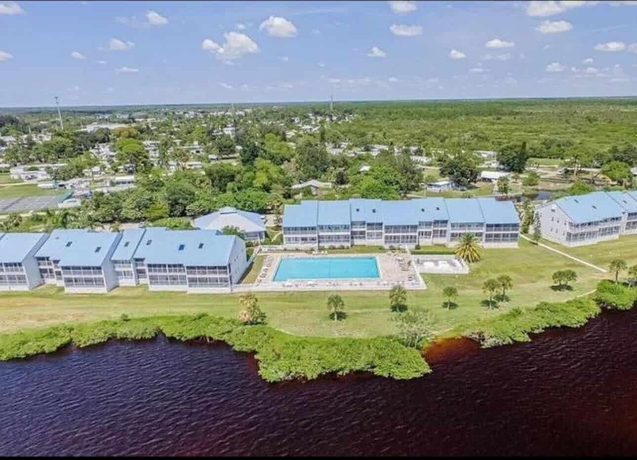 Aerial view of a residential complex with multiple buildings surrounding a large outdoor swimming pool, adjacent to a brownish river, with greenery and a small town in the background.