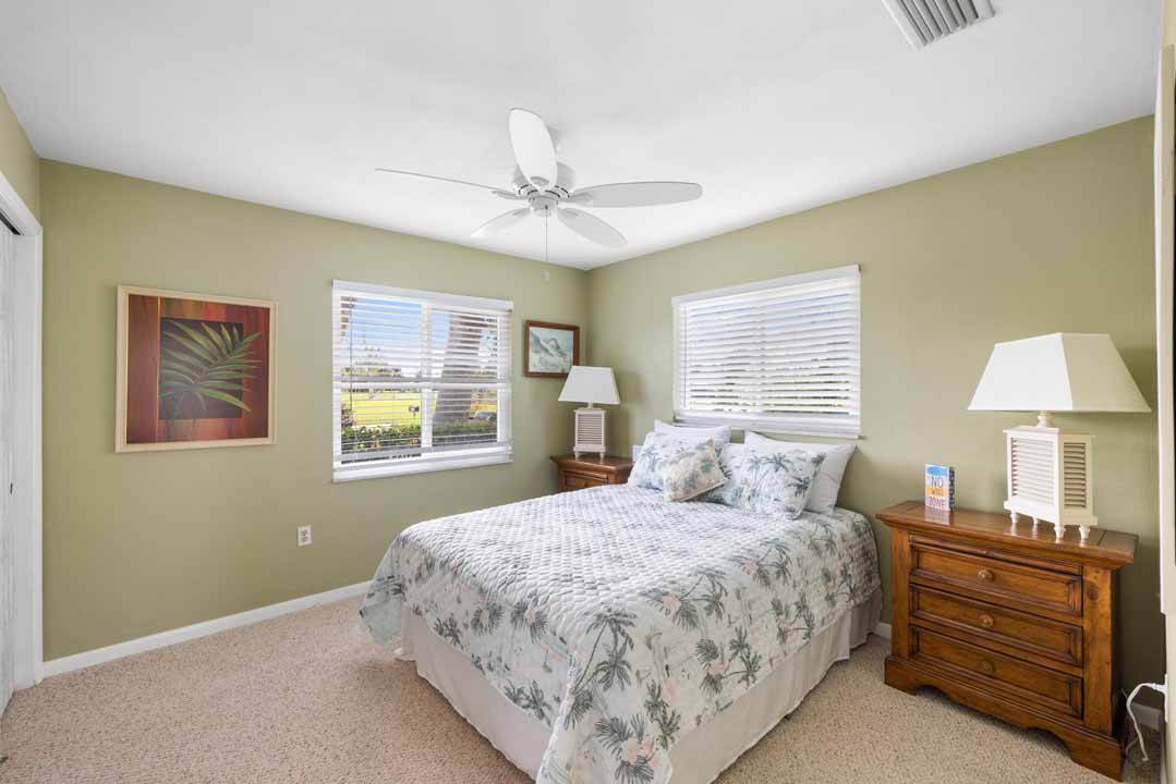 A well-lit bedroom in Rotonda, Florida, featuring a tropical theme with a bed adorned with floral bedding, wooden furniture, and a ceiling fan, offering views of the outdoors through two windows.